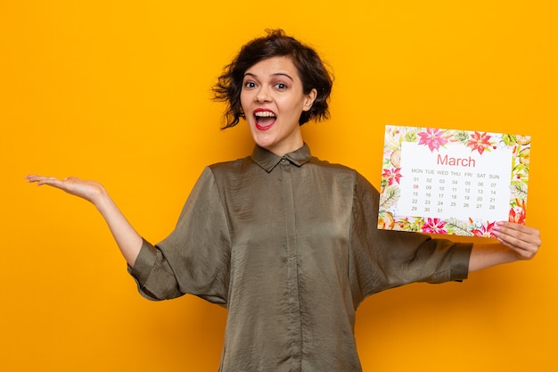 Foto donna felice con i capelli corti che tiene il calendario cartaceo del mese di marzo che sorride allegramente presentando con il braccio della mano che celebra la giornata internazionale della donna l'8 marzo in piedi su sfondo arancione