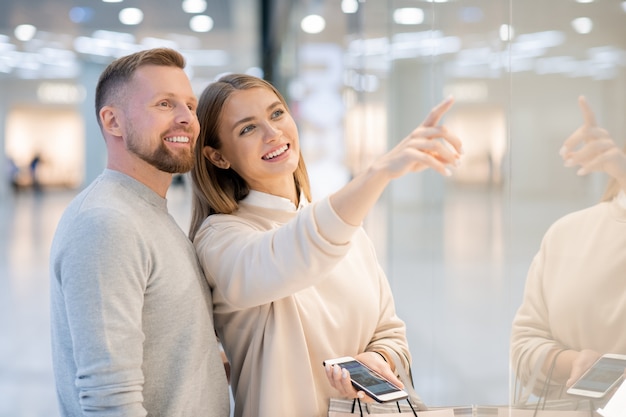 Счастливая женщина со смартфоном и бумажными пакетами показывает мужу модную повседневную одежду, указывая на витрину магазина