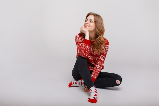 Happy woman with a smile in a red sweater with a print sits on the floor in a studio on a gray background