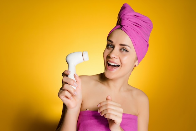 Una donna felice con un asciugamano in testa dopo la doccia tiene in mano una spazzola per una pulizia profonda della pelle del viso