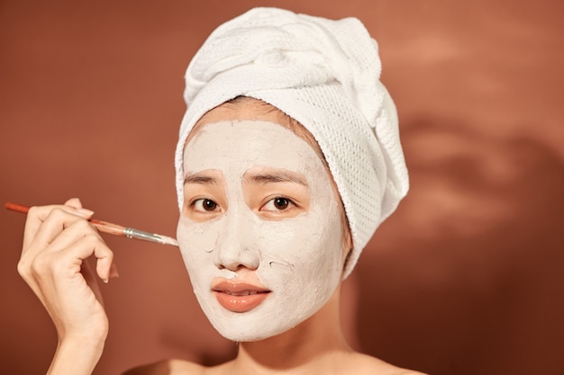 Счастливая женщина с полотенцем на голове наносит очищающую маску на лицо
