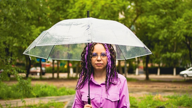 Happy woman with umbrella Cheerful female in a purple shirt holding umbrella and looking at camera while standing in green park on rainy day