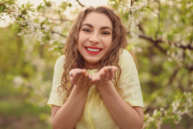 Photo happy woman with wavy hair holding hands like bowl and smiling for camera while standing on blurred background of blooming trees in spring garden