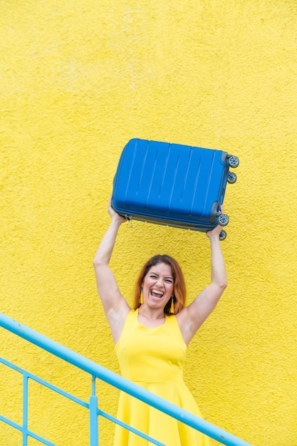 Una donna felice con un vestito giallo tiene una valigia blu sopra la testa sullo sfondo di un muro giallo la ragazza caucasica sorride con i denti e aspetta felicemente un viaggio