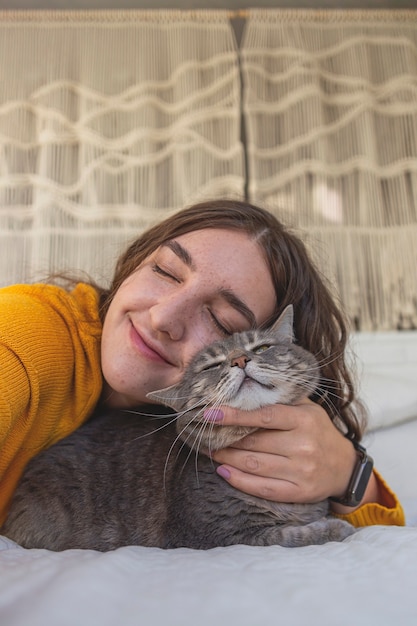 Happy woman in yellow sweater hugs her gray cat on the bed in a light interior