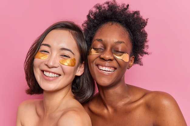 Happy women friends apply golden collagen patches under eyes to remove dark circles being in good mood stand shirtless have different skin condition