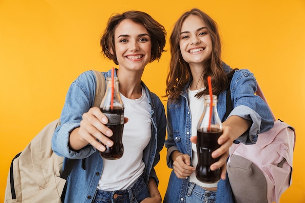 Happy women friends isolated over yellow wall drinking soda.