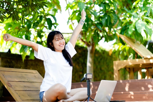 Счастлива работать Веселая молодая азиатская женщина улыбается, работая на своем ноутбуке за пределами офиса дома, работая в парке, работая, отдыхая дома и работая в Интернете