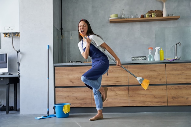 Happy worker full length shot of cheerful young woman cleaning lady pretending to sing a