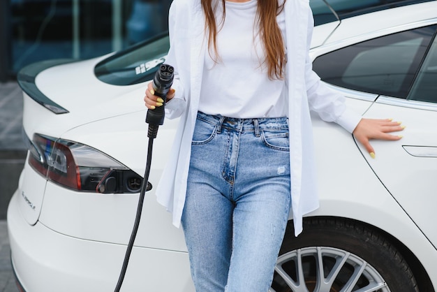 Happy young adult girl holding power cable supply in hand\
standing near electric car public charging station