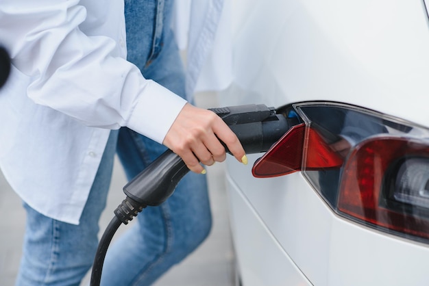 Happy young adult girl holding power cable supply in hand\
standing near electric car public charging station