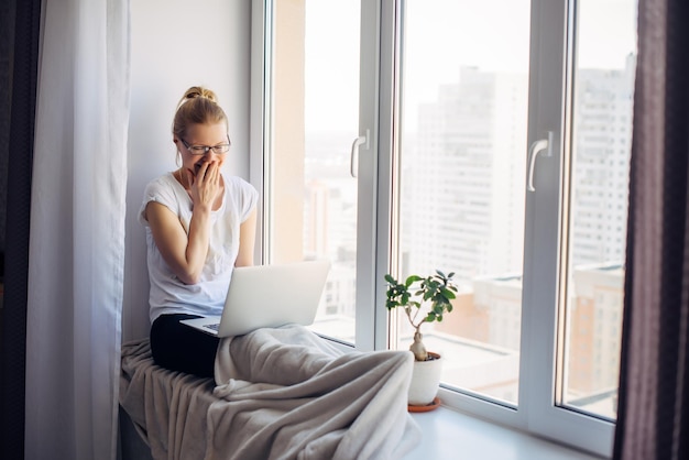 Happy young adult woman freelancer wearing glasses sitting on window background Laughing blonde working on laptop Workplace on the windowsill Home office concept