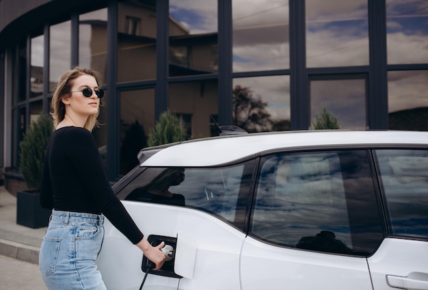 Happy young adult woman smiling wide looking away charging automobile battery from small public station standing near electric car drinking coffee and talking on smartphone