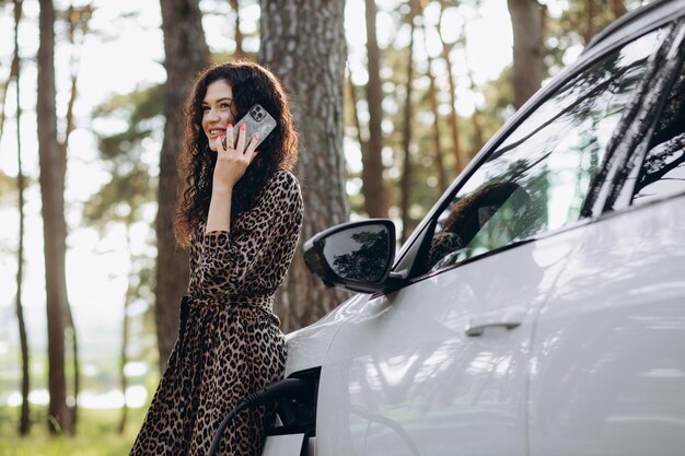 Happy young adult woman smiling wide looking away charging\
automobile battery from small public station standing near electric\
car drinking coffee and talking on smartphone