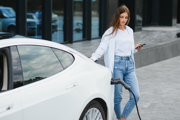 Foto felice giovane donna adulta sorridente distoglie lo sguardo caricando la batteria dell'automobile dalla piccola stazione pubblica in piedi vicino all'auto elettrica