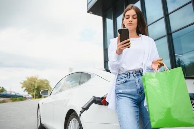 Happy young adult woman smiling wide looking away charging automobile battery from small public station standing near electric car