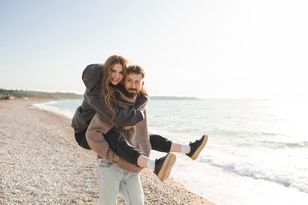 Happy young adults couple having fun at beach over sea background together Smiling man carry pretty woman over ocean Relationships