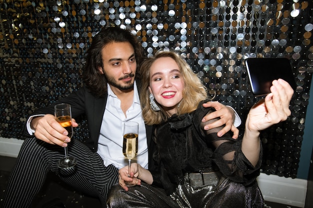 Happy young affectionate couple in glamorous attire making selfie by glittering wall in night club at party