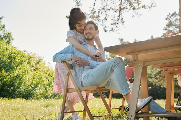 Happy young affectionate woman with glass of red wine embracing her smiling boyfriend sitting on chair by served table outdoors