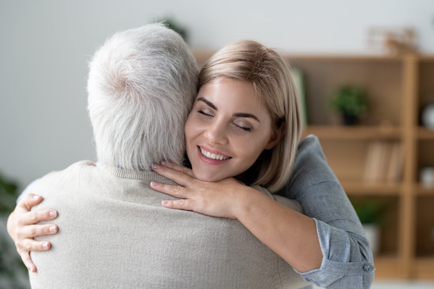 Happy young affectionate woman with toothy smile giving hug to her senior father while expressing love and care