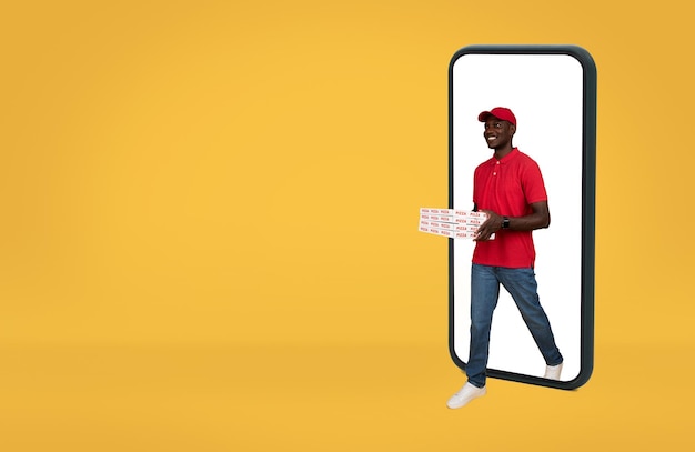 Photo happy young african american delivery man in uniform coming out of smartphone with blank screen with