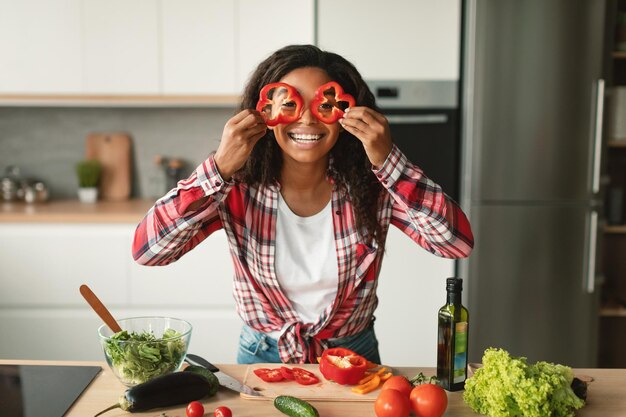 Foto la giovane donna afroamericana felice si mette i pezzi di pepe negli occhi come occhiali e si diverte in cucina