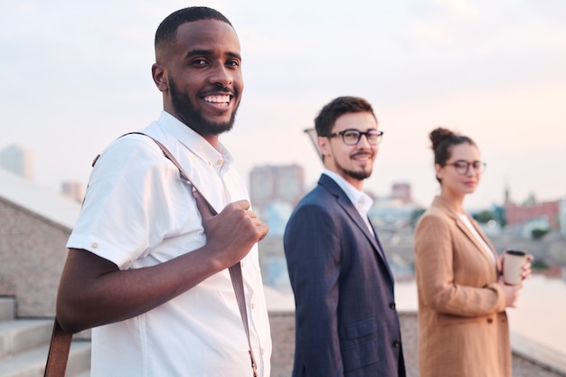 Happy young African businessman in smart casualwear looking at you while standing in front of camera against his colleagues outdoors