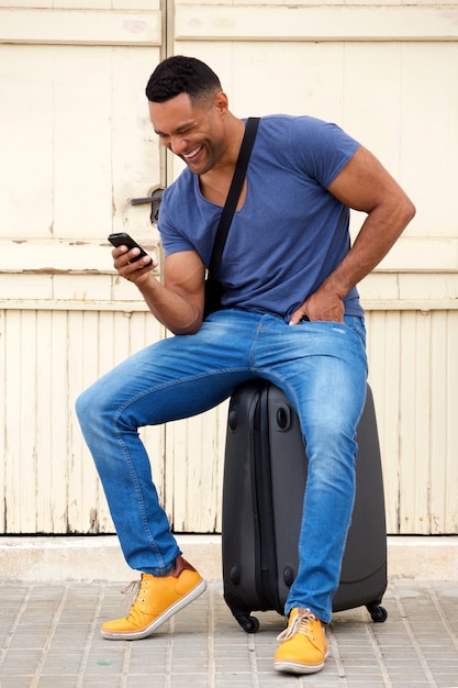 Happy young african man sitting on suitcase and using cell phone