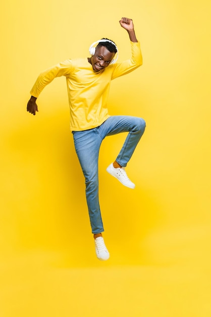 happy young african man wearing headphones listening to music and jumping with hand up in yellow isolated studio background