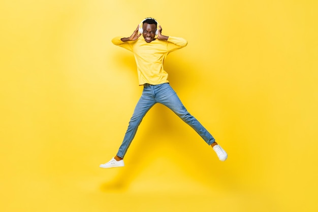 Happy young African man wearing headphones listening to music and jumping in yellow isolated studio background