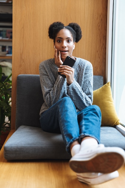 Ragazza felice giovane studente africano che studia presso la biblioteca, ascoltando musica con gli auricolari