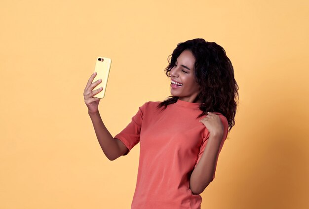 黄色の背景に携帯電話で祝う幸せな若いアフリカ女性selfie