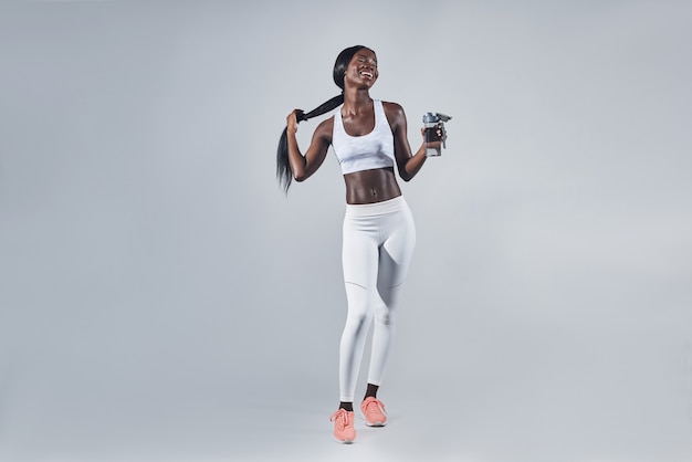 Happy young african woman in sports clothing holding bottle with water and adjusting hair