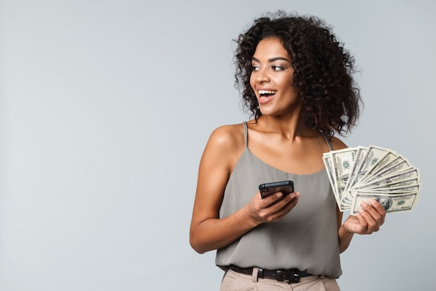 Photo happy young african woman standing isolated, holding bunch of money banknotes, using mobile phone