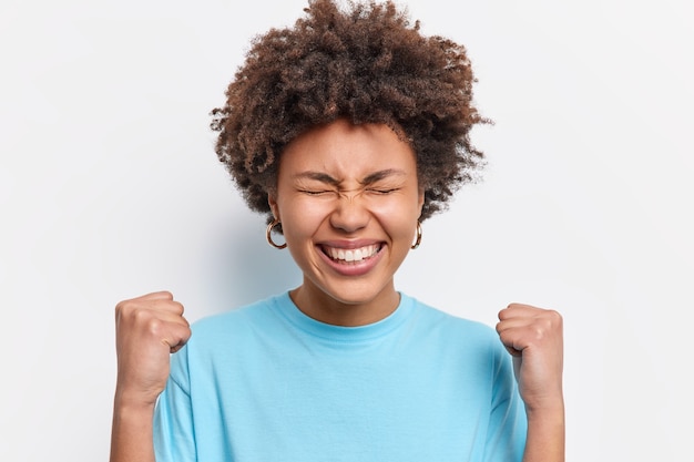 사진 행복 한 젊은 아프리카 계 미국인 여자 clenches 주먹 성공 미소에서 승리 승리를 축 하 광범위 하 게 식 흰 벽 위에 절연 캐주얼 파란색 t 셔츠를 착용하는 것을 기쁘게 생각합니다. 내가 이겼어