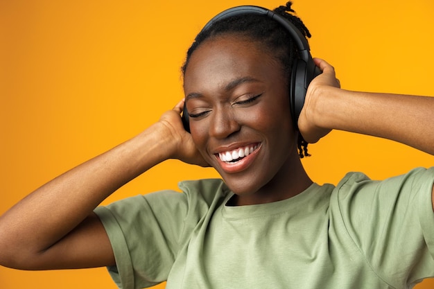 黄色の背景にヘッドフォンで音楽を聴いて幸せな若いアフロアメリカ人女性