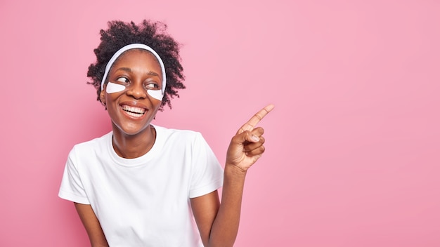 Happy young Afro American woman with curly hair smiles broadly shows white perfect teeth applies patches under eyes 