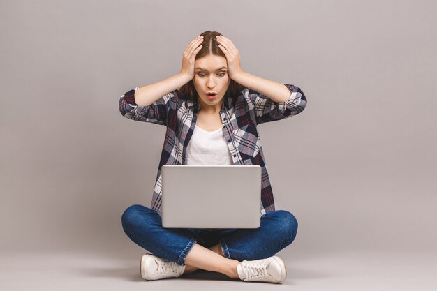 Happy young amazed woman sitting on the floor with crossed legs and using laptop 