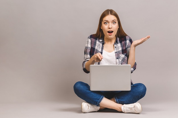 Happy young amazed woman sitting on the floor with crossed legs and using laptop