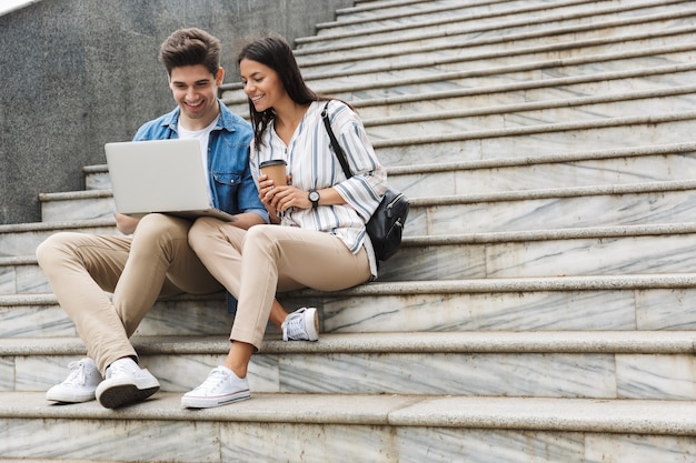 Felice giovane incredibile coppia amorevole uomini d'affari colleghi all'aperto sui gradini utilizzando il computer portatile a bere caffè.