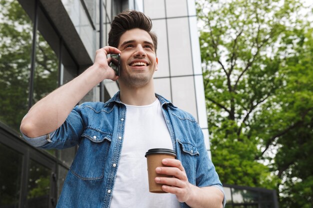 コーヒーを飲みながら携帯電話で話を歩いて外で屋外でポーズをとって幸せな若い驚くべき男のビジネスマン。