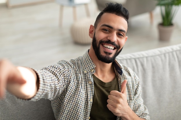 Happy young arab guy taking selfie and showing thumb up gesture\
smiling at mobile phone camera sitting on sofa at home