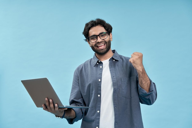 Happy young arab man celebrating win holding laptop isolated on blue