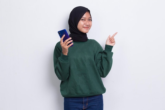 счастливая молодая азиатская красивая мусульманка с помощью мобильного телефона, указывающая в разные стороны