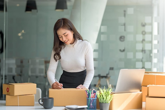 La giovane donna asiatica felice dell'imprenditore prepara la scatola dei pacchi e controlla gli ordini online del prodotto per la consegna al cliente sul computer portatile concetto di shopping online