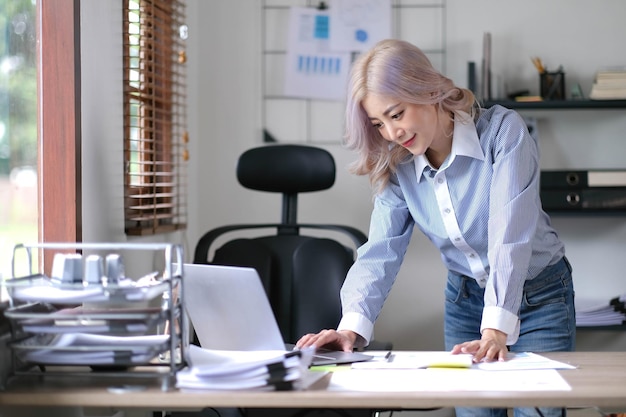 Happy young Asian businesswoman standing using calculator and laptop computer at office