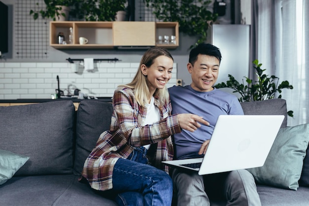 Счастливая молодая азиатская пара сближается, смеясь, используя ноутбук, сидя за столом дома