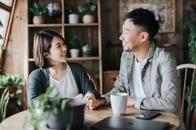 写真 カフェでコーヒーデートをしている幸せな若いアジア人カップルコーヒーテーブルで手をつないでコーヒーの飲み方とおしゃべりをし一緒にリラックスした瞬間を楽しんでいます