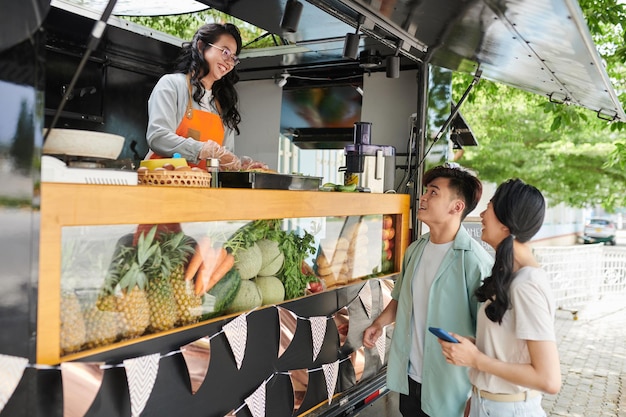 Happy young Asian couple standing by street food truck and looking at vendor