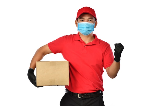 Happy young Asian delivery man in red uniform, medical face mask, protective gloves carry cardboard box in hands isolated on white. Delivery guy give parcel shipment. Safe delivery